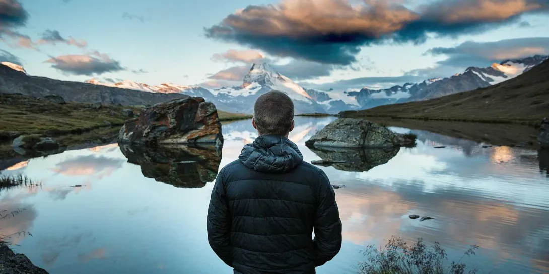Person in jacket gazes at lake reflecting mountains and clouds.